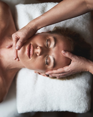 Massage Visage Relaxant : Soin Détente Profonde Femme aux yeux fermés recevant un massage relaxant du visage et des tempes sur une serviette blanche, gros plan vertical.