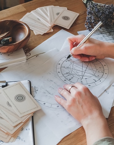 Thème Astral et Cartes de Tarot : Consultation Ésotérique Mains dessinant un thème astral complexe sur papier avec un stylo argenté, entouré de cartes de tarot et d'un bol en bois.